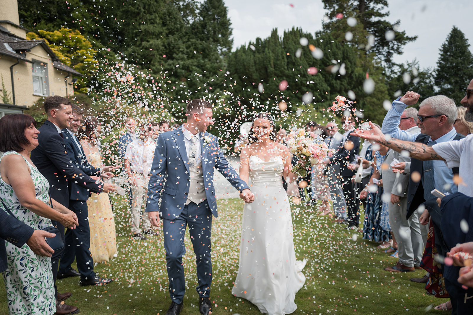 Alex and Dan walking through confetti surrounded by their guests at Lancrigg