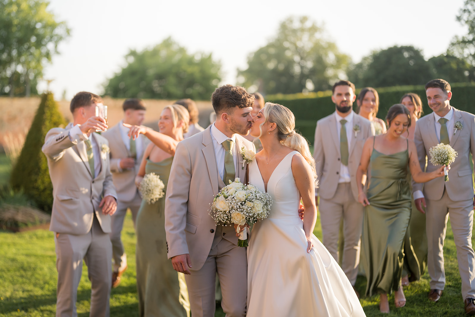 Bride and groom kissing with their wedding party in the gardens at Combermere Abbey
