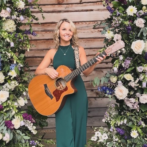 Daisy Kate with her acoustic guitar beside a floral arch
