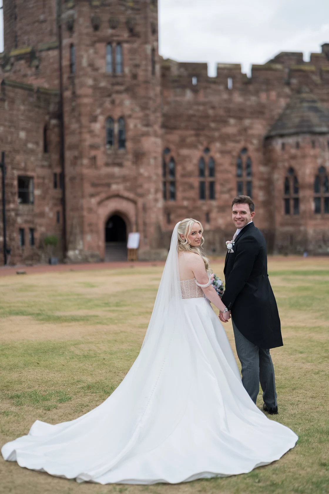 Hannah and Zak holding hands on the lawn in front of Peckforton Castle