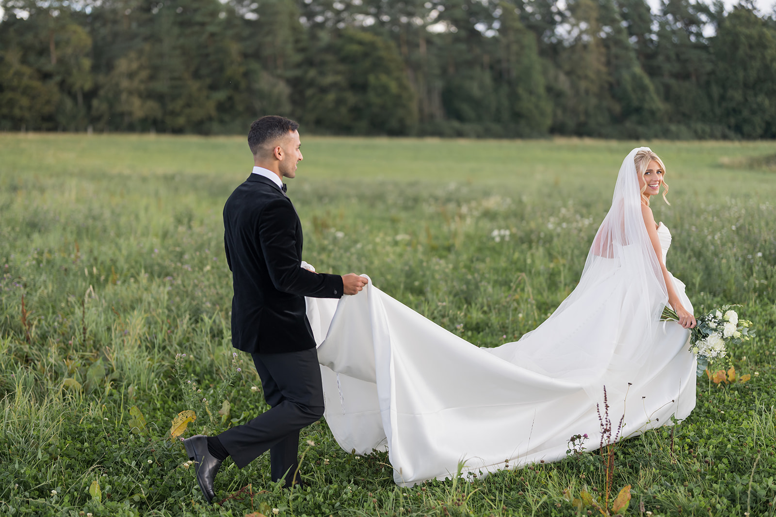 Groom holding the bride’s flowing veil in a meadow